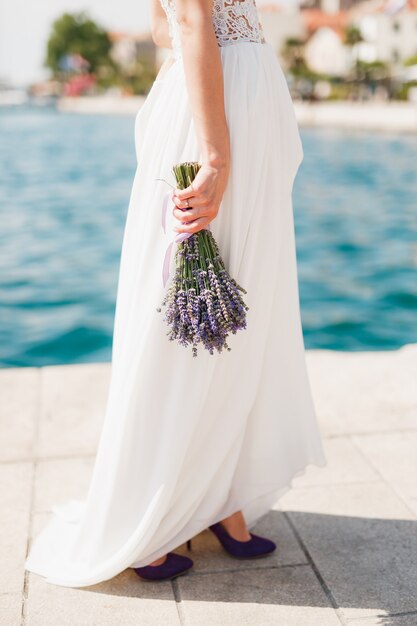 La mariée vêtue d'une robe en dentelle blanche avec un bouquet de lavande dans les mains se dresse sur la jetée au bord de la mer