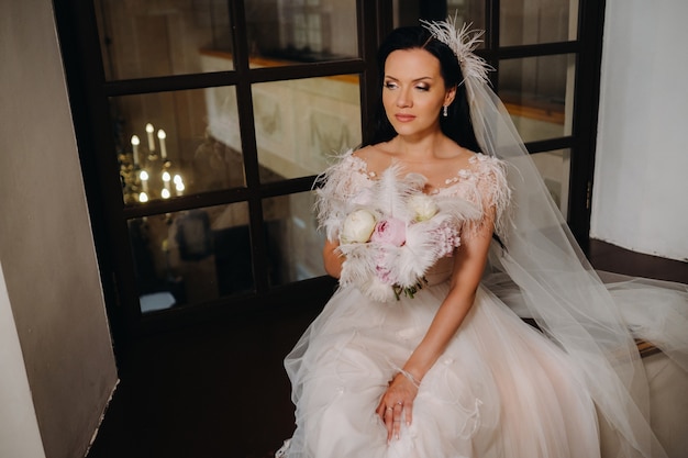 Une mariée vêtue d'une robe de mariée et d'un bouquet est assise devant une vieille fenêtre ouverte et regarde