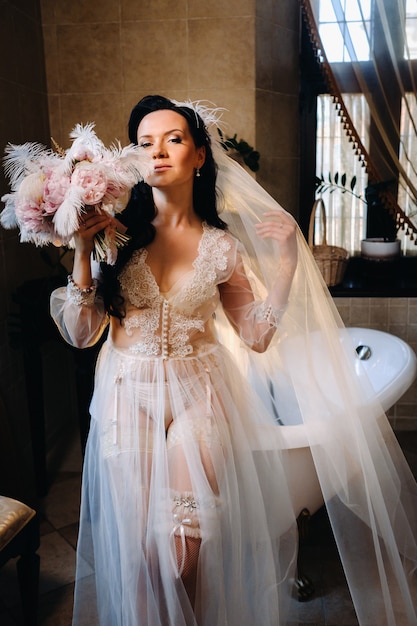 La mariée, vêtue d'une robe transparente boudoir et de sous-vêtements, est assise près d'un bain vintage avec une plume dans les mains, Le matin de la mariée.