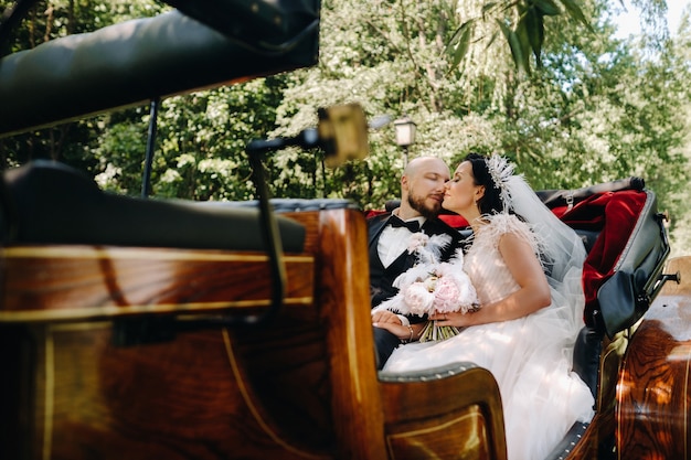 Les mariés avec un bouquet sont assis dans une calèche dans la nature dans un style rétro