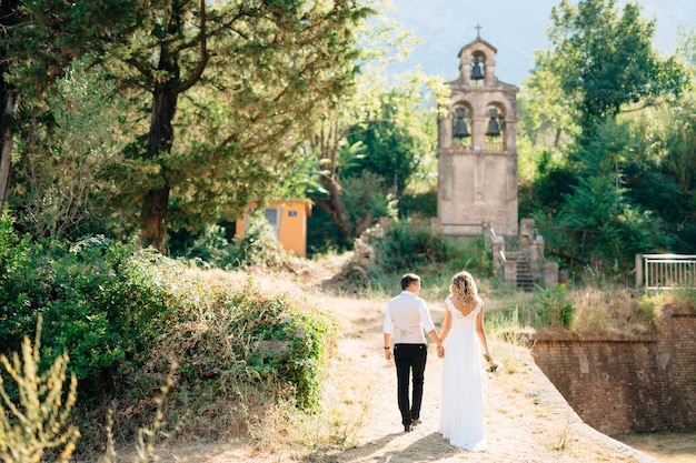 Les mariés marchent le long de la route illuminée vers l'ancien clocher se tenant la main