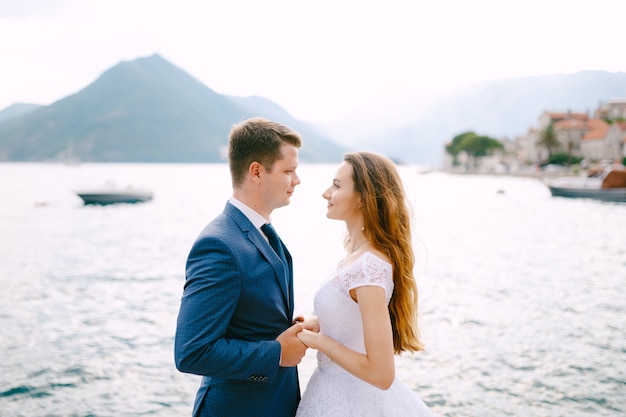 Les mariés se tiennent la main sur la jetée de Perast, derrière eux un bateau navigue