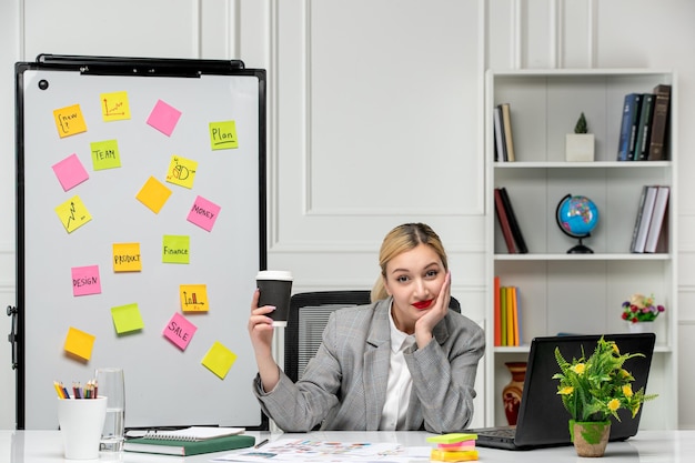 Marketing jeune jolie fille blonde en costume gris au bureau tenant la tête avec le visage et le café