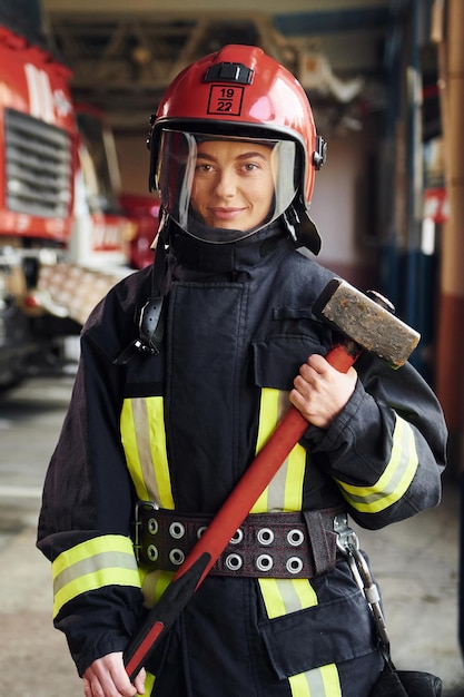 Avec un marteau dans les mains Femme pompier en uniforme de protection debout près du camion