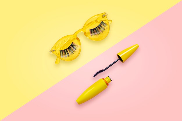 Mascara en tube jaune avec pinceau ouvert sur des lunettes de soleil roses et jaunes avec de faux cils.