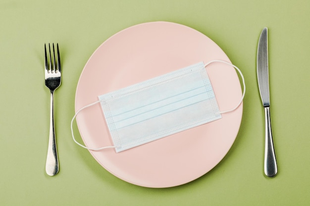 Masque sur une assiette vide avec une fourchette et un couteau sur fond vert Le concept de nutrition et de protection contre le virus