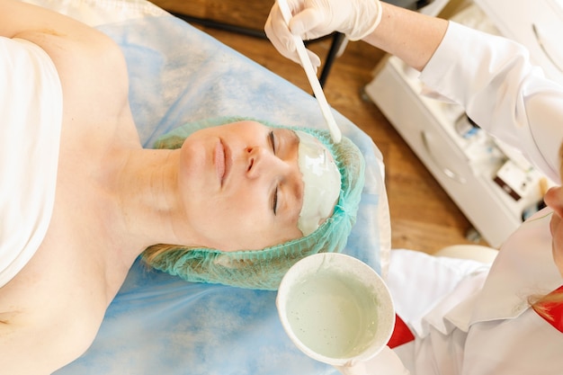 Masque facial au collagène. Traitement de la peau du visage. Femme recevant une procédure cosmétique dans un salon de beauté. Bouteille avec crème hydratante sur fond.