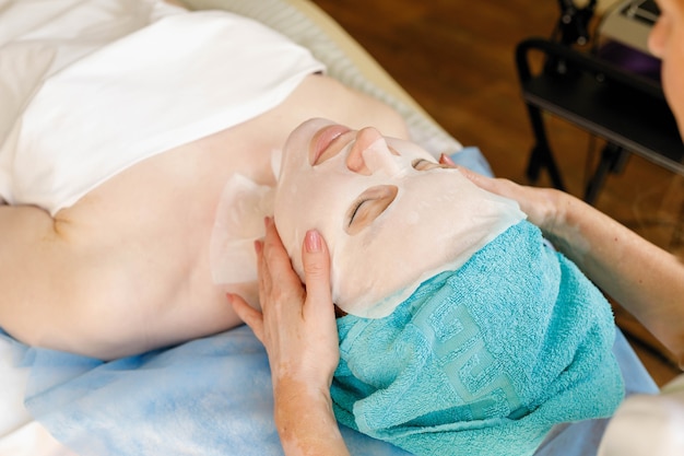 Masque facial au collagène. Traitement de la peau du visage. Femme recevant une procédure cosmétique dans un salon de beauté. Bouteille avec crème hydratante sur fond.