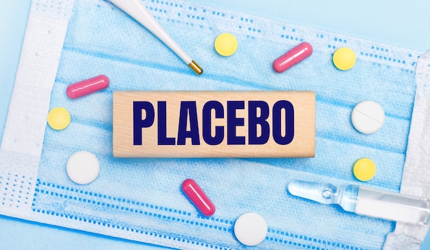 Sur Un Masque Facial Jetable Bleu Clair, Il Y A Des Comprimés, Un Thermomètre, Une Ampoule Et Un Bloc De Bois Avec Le Texte Placebo. Notion Médicale