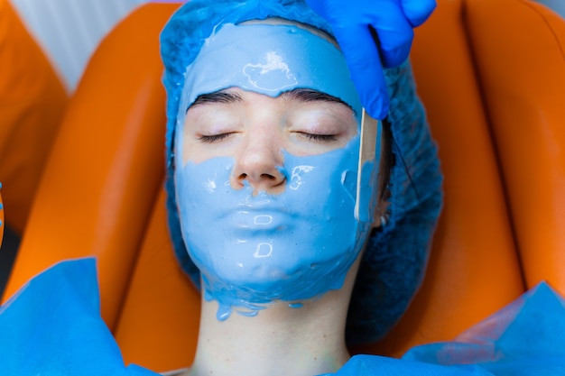 Masque hydratant à l'alginate pour le visage et la peau de la jeune fille. Procédure de spa pour le rajeunissement. esthéticienne frotte le masque bleu. Dermatologie en clinique médicale