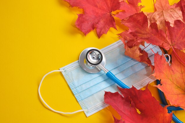 Masque médical de protection jetable et stéthoscope et feuilles d'érable d'automne sur fond jaune