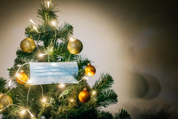 Masque de protection médical suspendu à un arbre de Noël décoré de boules brillantes et d'une guirlande lumineuse. Célébrer les vacances d'hiver en toute sécurité pendant la pandémie de coronavirus. Concept de covid et bonne année.