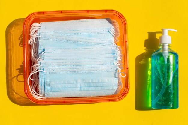 Masques bleus avec désinfectant en gel liquide alcool dans une boîte rouge sur une surface jaune