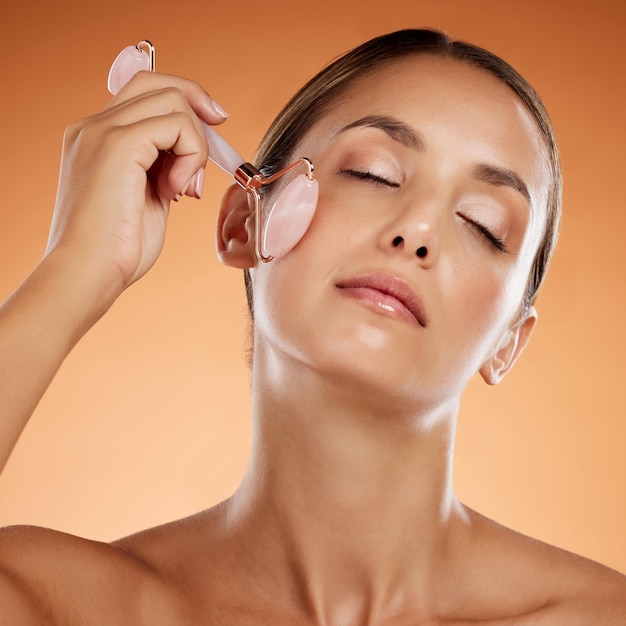 Massage du visage de la beauté de la femme et rouleau de quartz rose produit cosmétique anti-âge et outil de soin esthétique sur fond de studio orange