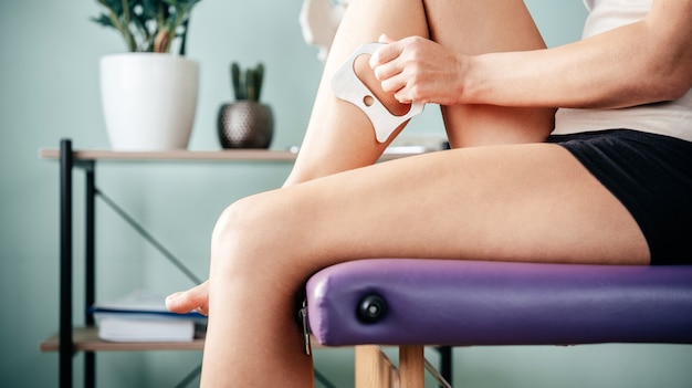 Massage Guasha sefl de la jambe à l'aide de l'outil IASTM pour le traitement de la restriction myofasciale, une femme est assise sur une table de massage et se gratte la jambe avec l'outil guasha IASTM