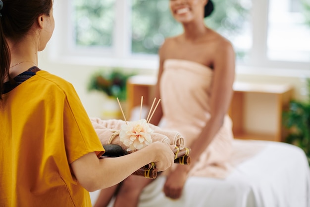 Masseur apportant un plateau avec des serviettes roulées à chaud à smiling young woman sitting on bed in spa salon