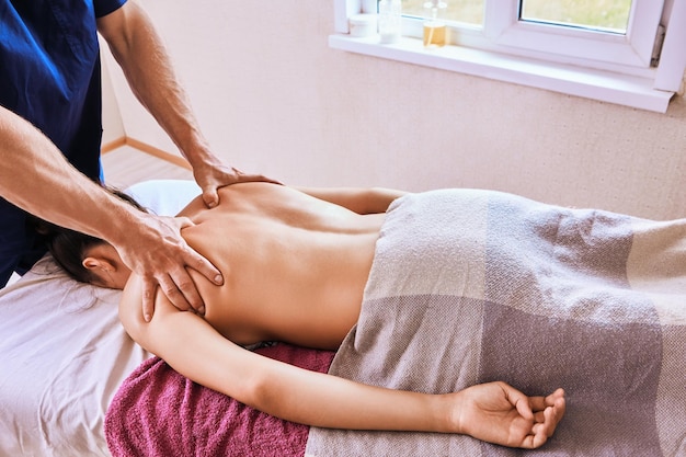 Le masseur fait un massage profond des tissus sur la zone de l'épaule d'une jeune femme blanche allongée face vers le bas pendant le massage