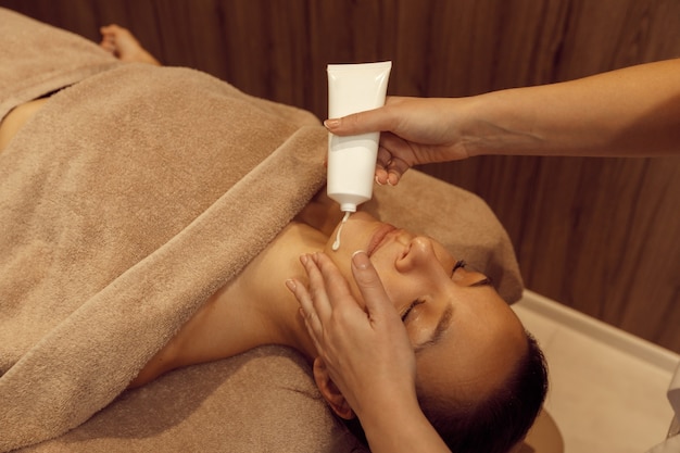 Une masseuse applique de la crème sur le visage de la femme, un massage professionnel. Massages et relaxation, soins du corps et de la peau. Dame attirante dans le salon de station thermale