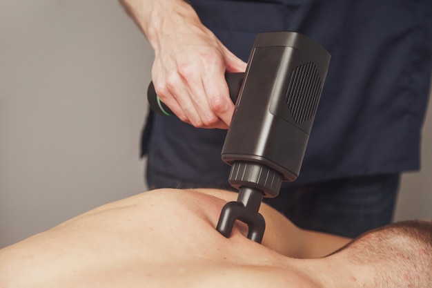 Le massothérapeute traite les blessures du patient masculin athlète professionnel. Massages de choc au pistolet sportif dans le cabinet médical de la salle de sport. Thérapie par percussion pour le massage de régénération du corps athlétique. Physiothérapie