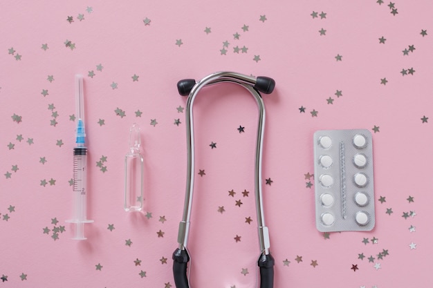 Matériel de médecine stéthoscope ou phonendoscope, blister, seringue et ampoule transparente en verre sur fond rose avec des étoiles de confettis.