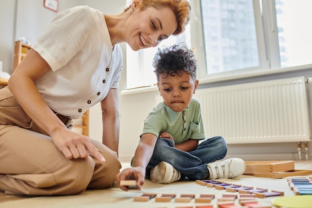 Photo matériel montessori garçon afro-américain intelligent jouant à un jeu de couleur éducatif près de l'enseignant heureux