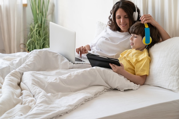 Matin en famille le week-end maman travaille sur un ordinateur portable allongé dans son lit avec un enfant de tenir la tablette jouer à des jeux