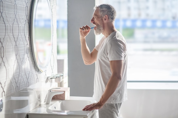 Matin, Lavage. Homme Barbu Aux Cheveux Gris En Tshirt Blanc Se Brosser Les Dents Devant Le Miroir De Bonne Humeur