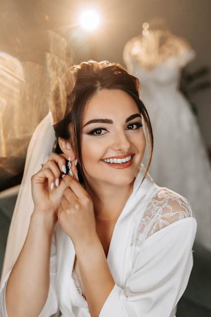Photo le matin de la mariée dans une robe blanche et une coiffure élégante portrait d'une fille attachant une boucle d'oreille regardant dans l'objectif de la caméra et souriant