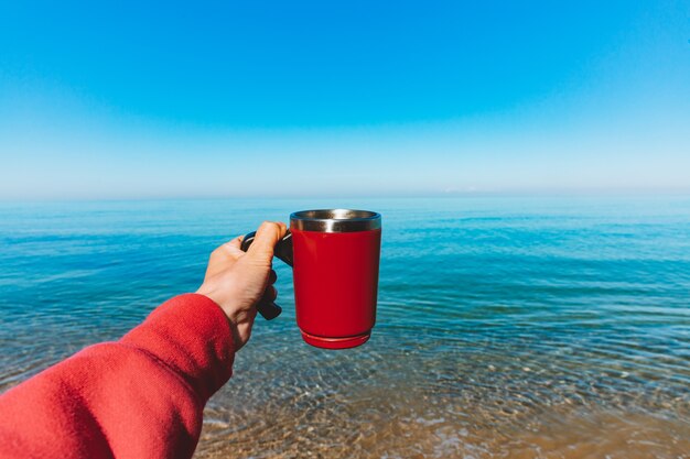 Matin en mer. La fille tient une tasse thermo rouge avec du café ou du thé chaud. Vacances d'été dans la nature Main close up Ustensiles de camping