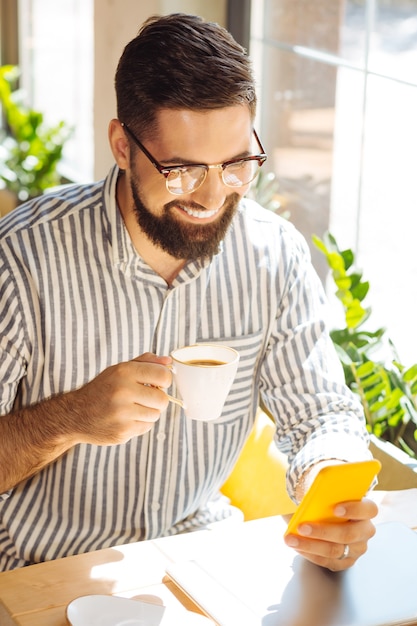 Matin parfait. Heureux homme ravi utilisant son smartphone tout en savourant sa tasse de café au café