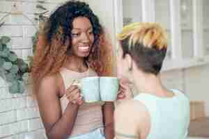 Photo matinée réussie. femme heureuse à la peau sombre avec de longs cheveux bouclés avec café et petite amie avec dos à la caméra communiquant dans la cuisine