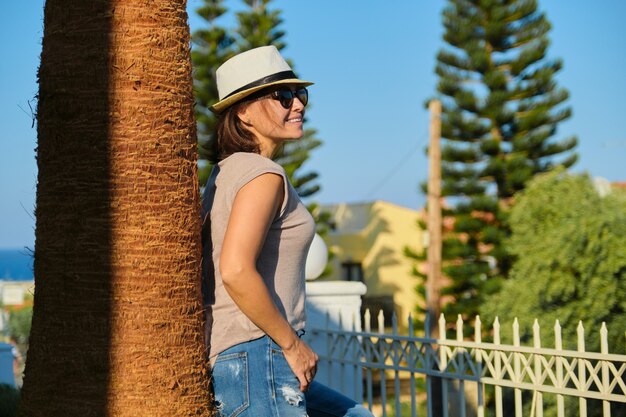 Mature heureuse belle femme en chapeau de lunettes de soleil debout près de palmier au coucher du soleil