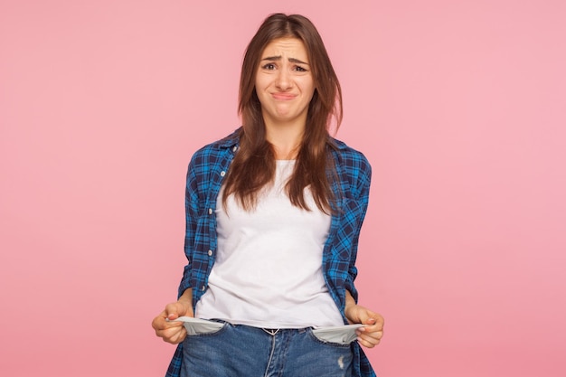 Mauvais étudiant, chômage. Portrait d'une jeune fille sans emploi bouleversée en chemise à carreaux qui sort des poches vides, ne montrant aucun geste d'argent, inquiète des dettes. studio d'intérieur tourné isolé sur fond rose