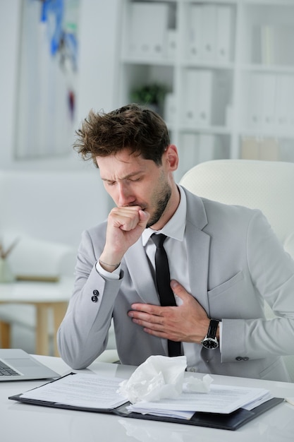 Mauvais homme d'affaires avec barbe assis au bureau avec des documents et tenant la main sur la poitrine tout en ayant des symptômes de toux, de coronavirus ou de pneumonie