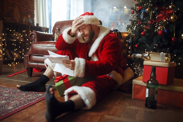 Mauvais père noël ivre lit des lettres sous l'arbre de Noël