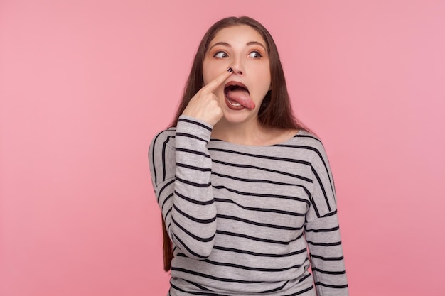 Mauvaise habitude Portrait d'une femme drôle en sweat-shirt rayé forant le nez avec une expression stupide humoristique faisant une grimace stupide qui sort la langue prise de vue en studio à l'intérieur isolée sur fond rose