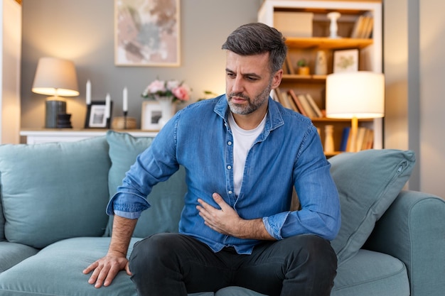 Photo maux d'estomac terribles jeune homme beau frustré étreignant son ventre et gardant les yeux fermés tout en étant allongé sur le canapé à la maison