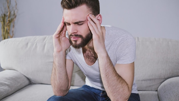 Maux de tête ou migraine Un jeune homme barbu tient ses mains derrière sa tête tout en étant assis sur le canapé à la maison