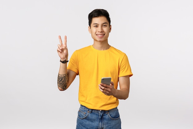 Mec asiatique hipster mignon et gai avec des tatouages, montrant le signe de la paix et tenant le téléphone, souriant joyeux, messagerie, utilisant une application, envoyant la positivité tout au long des blogs, mur blanc