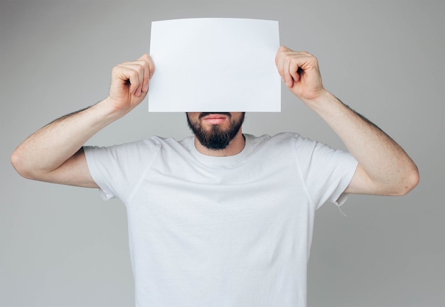 Un mec barbu couvre une partie du visage avec une page blanche blanche. Tenez-le à deux mains