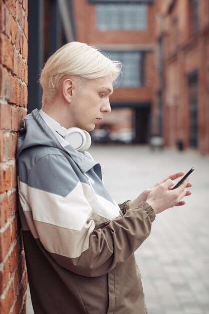 Mec blond avec des écouteurs sur son cou faisant défiler la liste de lecture dans le smartphone