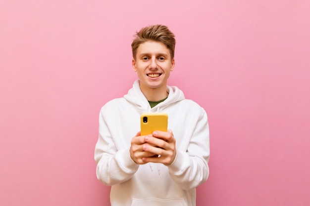Un mec blond en vêtements décontractés blancs se dresse sur un fond rose avec un smartphone dans les mains