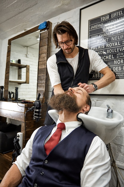 Mec brutal dans un salon de coiffure moderne. Le coiffeur fait de la coiffure un homme avec une longue barbe. Le coiffeur principal fait la coiffure avec des ciseaux et un peigne