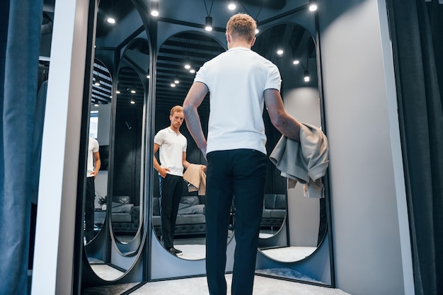 Un mec en chemise blanche essaie de nouveaux vêtements et se regarde dans le miroir