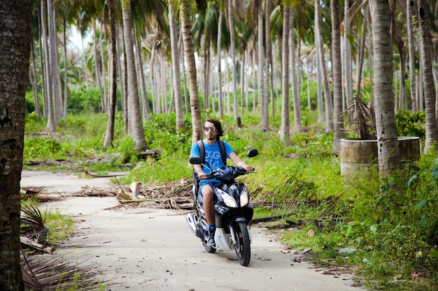 Un mec en chemise bleue conduit une moto dans la jungle parmi les palmiers