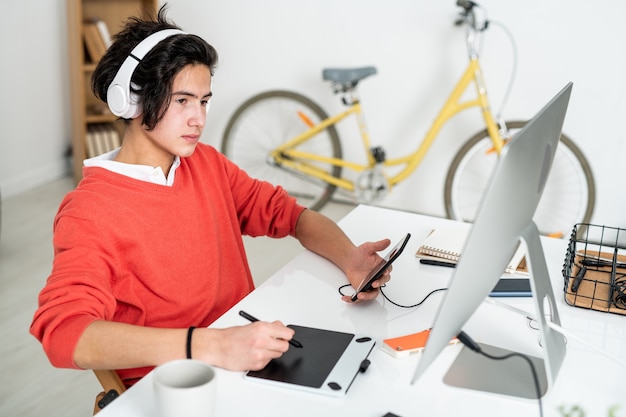 Mec contemporain avec des écouteurs assis par un bureau, à l'aide d'un smartphone, regardant un écran d'ordinateur et retoucher des photos