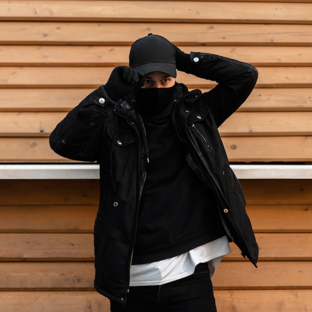 Un mec cool et élégant avec un masque de protection noir dans des vêtements de mode noirs porte une casquette noire près d'un mur en bois