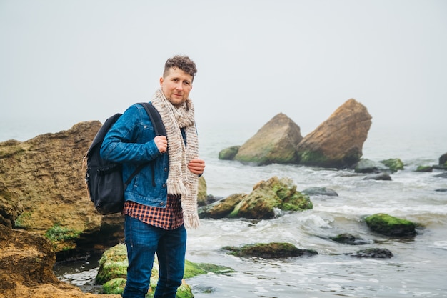 Mec hipster élégant avec un sac à dos debout sur des rochers sur fond de mer