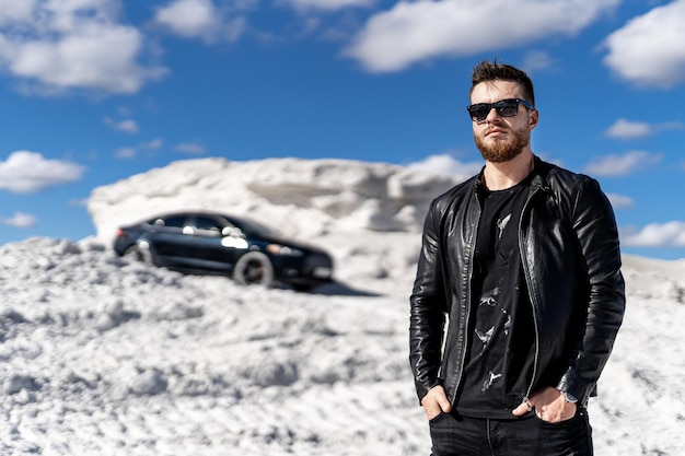 Mec mignon modèle de mode élégant sur photosession dans une carrière. Voiture de luxe en arrière-plan. homme riche en veste de cuir et lunettes de soleil.