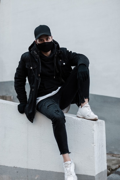 Mec à la mode dans des vêtements noirs élégants avec une casquette noire, une veste, un sweat à capuche, un jean et des baskets blanches avec un masque de protection assis à l'extérieur